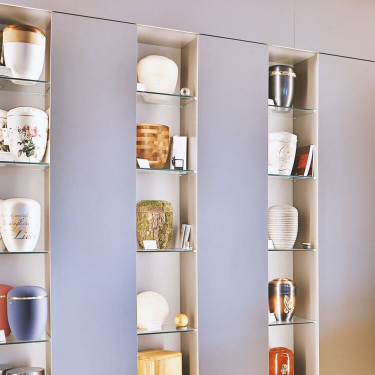 Mehrere Urnen in einem Ausstellungsraum in drei unterschiedlichen Regalen. Jede Urne steht einzeln in einem eigenen gut beleuchteten Fach.