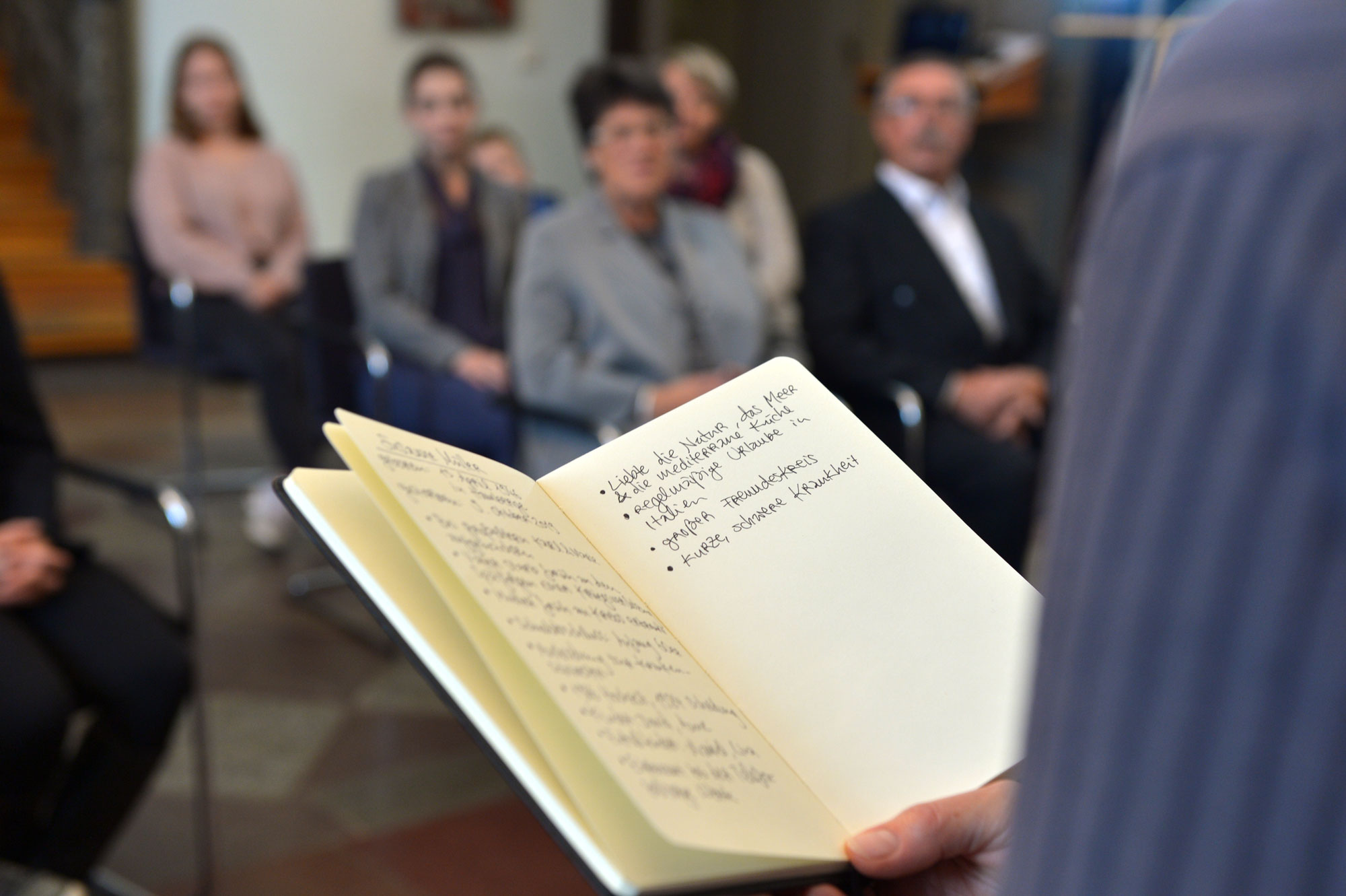 Sitzende Trauergemeinde im Hintergrund. Im Vordergrund hält ein Trauerredner ein Buch mit Notizen über den Verstorbenen in der Hand.