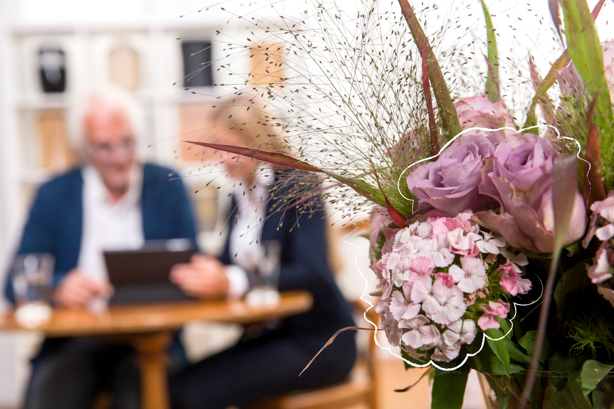 Zwei Personen schauen auf ein Tablet. Im Vordergrund verdeckt ein Blumenstrauß die Personen leicht.